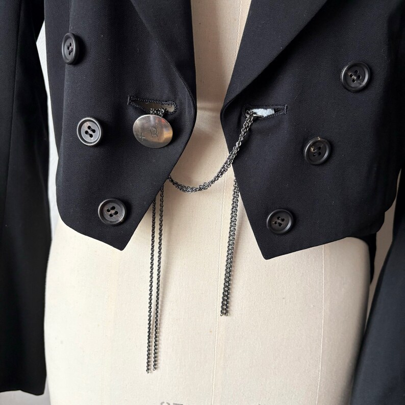 May include: A black cropped blazer with a silver chain and a circular pendant. The blazer has multiple buttons and a pointed collar.