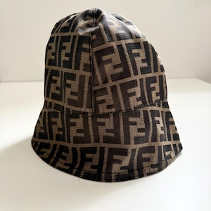 May include: A brown and black bucket hat with a repeating pattern of the letter "F". The hat has a rounded crown and a slightly downward-sloping brim. The fabric appears to be a woven material.