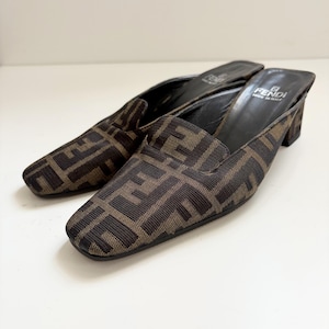 May include: A pair of brown and black Fendi mules. The shoes have a repeating geometric pattern and a small block heel. The interior of the shoe is black leather, and the Fendi logo is visible on the insole.
