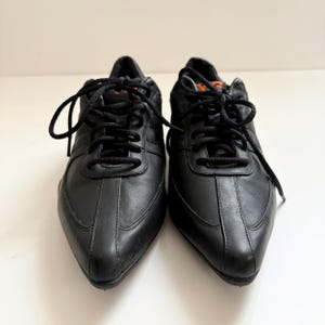 May include: A pair of black leather sneakers with a pointed toe design, featuring black laces and a small orange Y-3 logo on the tongue. The shoes have a smooth finish, a low-top silhouette, and a structured construction with visible stitching details.