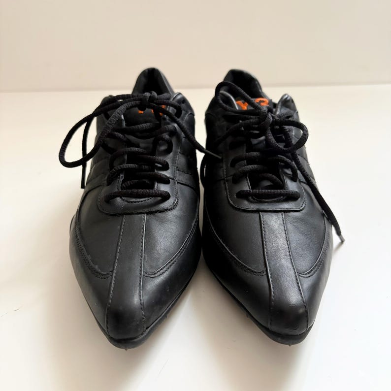 May include: A pair of black leather sneakers with a pointed toe design, featuring black laces and a small orange Y-3 logo on the tongue. The shoes have a smooth finish, a low-top silhouette, and a structured construction with visible stitching details.