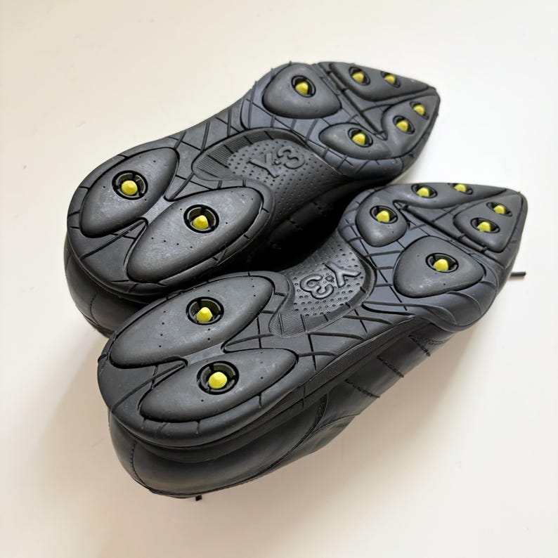 May include: A pair of black athletic shoes featuring a textured rubber outsole with a grid pattern and multiple bright yellow circular traction studs. The Y-3 logo is embossed in the center of the sole, showcasing a sleek, functional design.