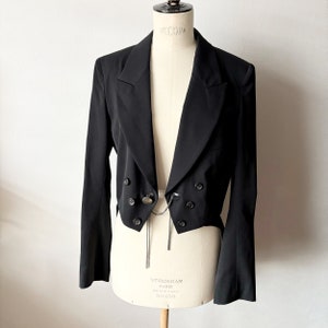 May include: A black cropped blazer with a double-breasted closure and a long chain detail. The blazer has a pointed collar and long sleeves.