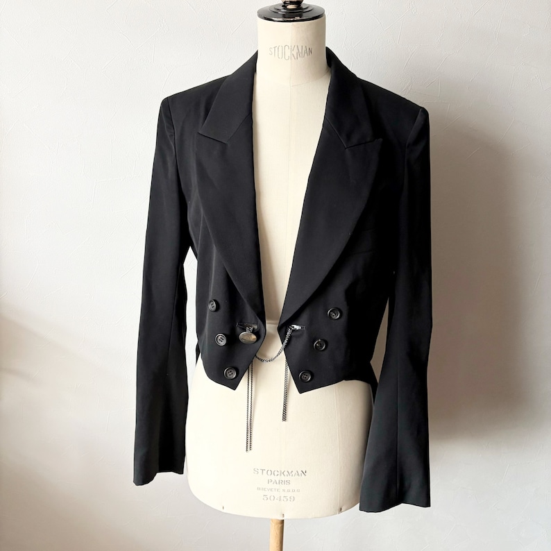 May include: A black cropped blazer with a double-breasted closure and a long chain detail. The blazer has a pointed collar and long sleeves.