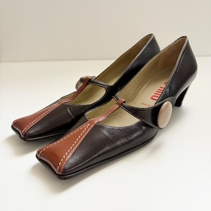 May include: A pair of dark brown leather Mary Jane heels with a square toe and a contrasting tan leather accent. The shoes feature a T-strap design and a small, rounded heel. White stitching details the edges.