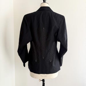 Puede incluir: Blazer negro con costuras decorativas blancas en la espalda, exhibido en un maniqu&iacute;. Las costuras forman un patr&oacute;n vertical. El blazer tiene un cuello cl&aacute;sico y mangas largas. El dise&ntilde;o a&ntilde;ade un toque distintivo a la prenda.