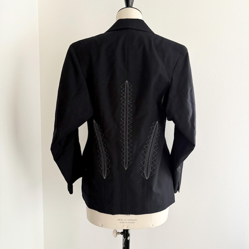Puede incluir: Blazer negro con costuras decorativas blancas en la espalda, exhibido en un maniqu&iacute;. Las costuras forman un patr&oacute;n vertical. El blazer tiene un cuello cl&aacute;sico y mangas largas. El dise&ntilde;o a&ntilde;ade un toque distintivo a la prenda.
