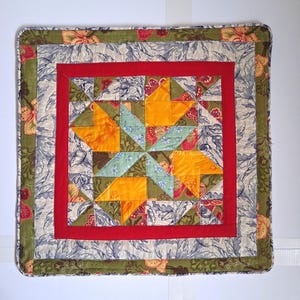 May include: A square, handmade quilt with a star pattern. The quilt features a red border around the star, with a floral and leaf pattern in the outer border. The star is made of orange, green, and blue fabrics.