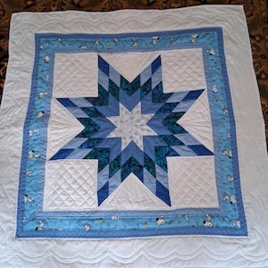 May include: A square quilt featuring a central eight-pointed star in shades of blue and teal, surrounded by a white quilted border and a light blue frame patterned with Snoopy characters and musical notes, finished with a white scalloped edge.