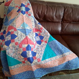 May include: A patchwork quilt featuring a geometric pattern with alternating blocks of solid blue, teal, and coral fabric alongside sections of floral-printed fabric in shades of blue, pink, and white, finished with visible machine-stitched quilting.