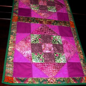 May include: A handmade quilted table runner featuring a patchwork design. The runner showcases a vibrant color palette of pinks, purples, and greens. The central design includes diamond and square patterns, with a green border. The runner is approximately 40 inches long.
