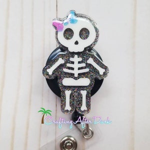 Pode incluir: Um carretel de crachá de glitter preto com uma figura de esqueleto branca usando um laço rosa e azul. O esqueleto é uma menina com uma cabeça de caveira e ossos longos. O carretel tem um porta-crachá de plástico transparente anexado. "Crafting After Dark" está escrito na parte inferior do carretel.