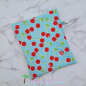 May include: A rectangular, light blue pouch with a repeating pattern of red cherries and green leaves. The pouch has a silver zipper and a small, circular logo. The text "Crafting After Dark" is visible at the bottom.
