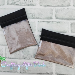 May include: Two clear zippered pouches with black fabric and a glittery gold background. The pouches are on a white brick background.