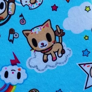 Puede incluir: Una tela azul claro con un patr&oacute;n repetido de un gato de dibujos animados con chispas en su pelaje, una dona con chispas, un arco&iacute;ris, una nube con una cara y estrellas. El gato sostiene un palo cubierto de chocolate.