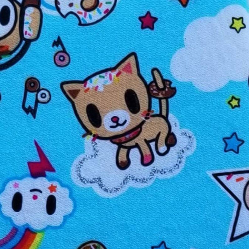 Puede incluir: Una tela azul claro con un patr&oacute;n repetido de un gato de dibujos animados con chispas en su pelaje, una dona con chispas, un arco&iacute;ris, una nube con una cara y estrellas. El gato sostiene un palo cubierto de chocolate.