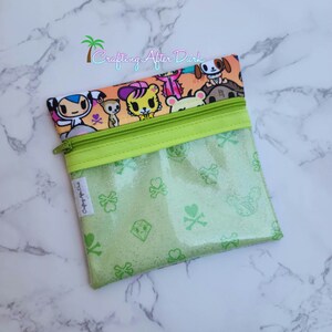 May include: A green and white zippered pouch with a clear front panel. The front panel has a green glitter background with a repeating pattern of skulls, hearts, and diamonds. The back panel has a colorful cartoon print of animals and the text "Crafting After Dark".