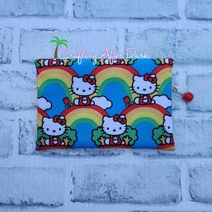 May include: A rectangular pouch featuring a repeating pattern of Hello Kitty characters, rainbows, and clouds against a blue background. The pouch has a red zipper pull with a small red bead. The text "Crafting After Dark" is printed on the top.