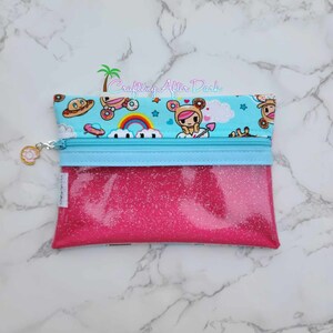 Puede incluir: Una bolsa rosa con purpurina con cremallera, con una cremallera azul y un forro de tela azul y blanco con un arco&iacute;ris, nubes y personajes de dibujos animados. La bolsa tiene un peque&ntilde;o amuleto unido a la cremallera.