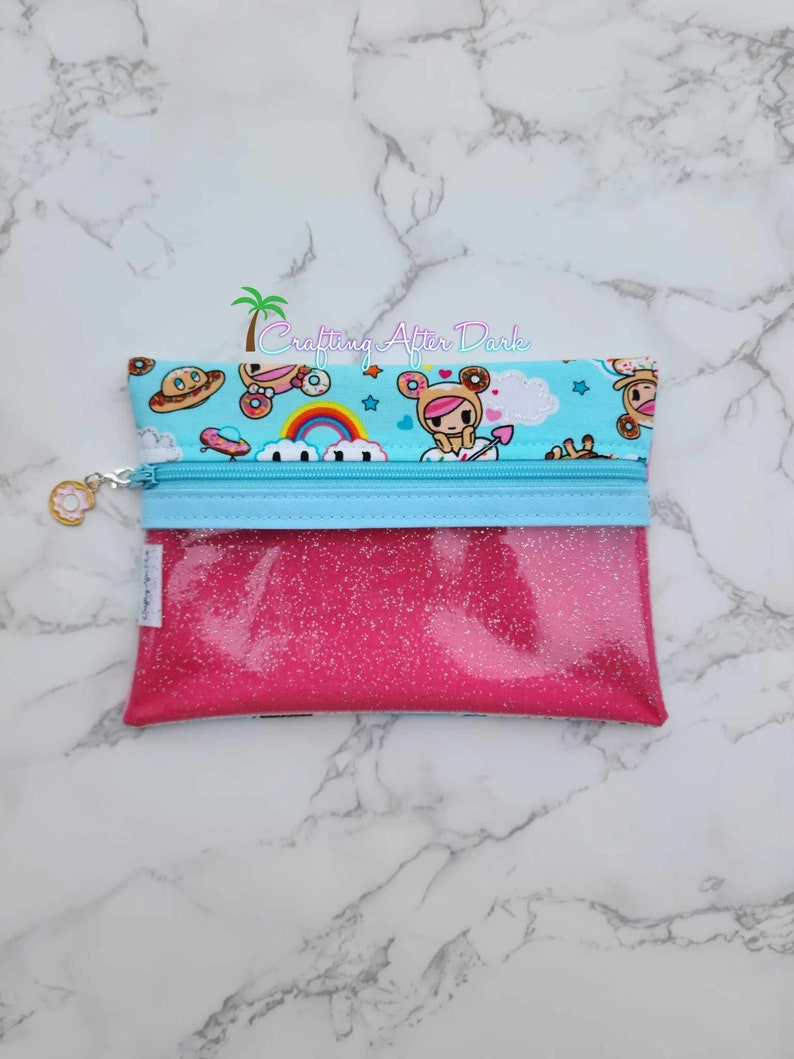 Puede incluir: Una bolsa rosa con purpurina con cremallera, con una cremallera azul y un forro de tela azul y blanco con un arco&iacute;ris, nubes y personajes de dibujos animados. La bolsa tiene un peque&ntilde;o amuleto unido a la cremallera.