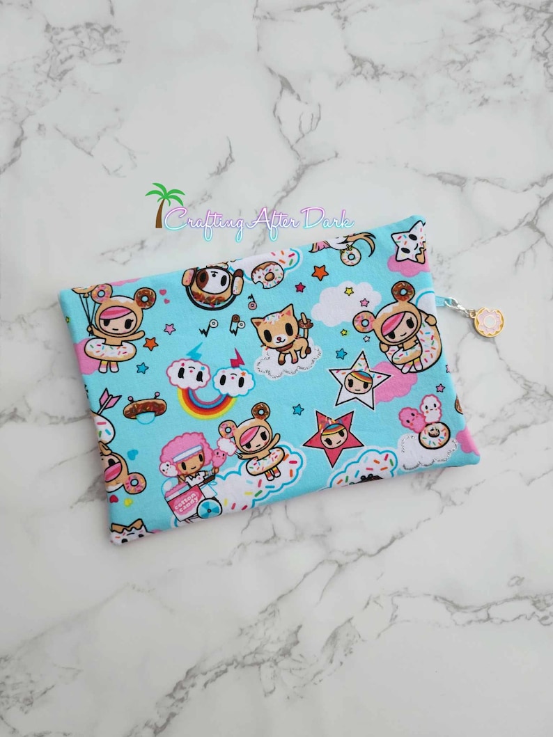 Puede incluir: Una bolsa azul con cremallera con un estampado de donuts y nubes con personajes de dibujos animados. La bolsa tiene un tirador de cremallera con forma de donut.