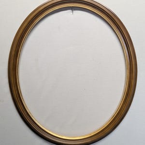 May include: Oval picture frame with a warm, golden-brown finish. The frame has a detailed, layered design and a small hook at the top for hanging. The frame is empty, ready for a photograph or artwork.
