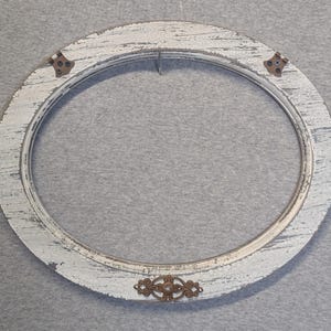 May include: An oval picture frame with a distressed white painted finish showing wood grain texture. The frame features two small bronze-colored metal corner brackets at the top and a decorative bronze-colored metal filigree accent at the bottom.