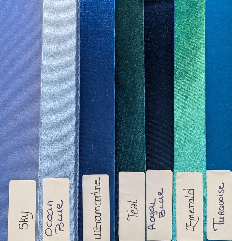 May include: A collection of seven vertical velvet ribbons in shades of sky, ocean blue, ultramarine, teal, royal blue, emerald, and turquoise. Each ribbon features a white rectangular label at the bottom with the corresponding color name written in ink.