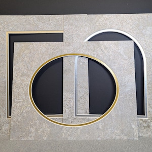 May include: A collection of decorative picture frames in various shapes. The frames feature a textured, light gray surface with gold and silver accents. The shapes include a rectangle, an oval, and an arch, with black backgrounds.