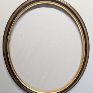 May include: Oval picture frame with a dark brown outer edge, a black inner edge, and a gold inner rim. The frame is empty, with a white background visible through the opening.