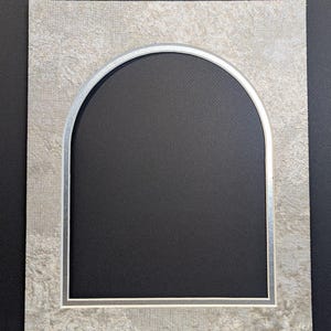 May include: A decorative picture mat with an arched opening. The mat has a textured, light gray surface with a silver border around the opening. The background is black.
