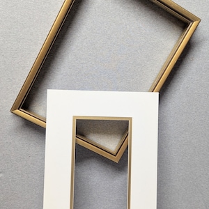 May include: Two gold-colored picture frames, one rectangular and one square, are positioned against a gray background. A white mat with a gold inner border is in the foreground. The frames appear to be empty.