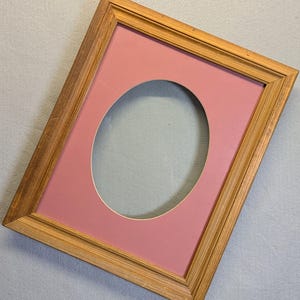 May include: A wooden picture frame with a pink mat and an oval cutout. The frame has a natural wood finish.