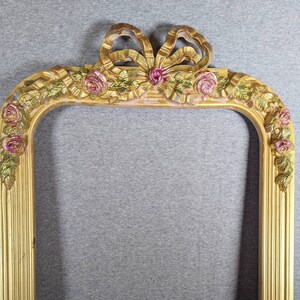 May include: Ornate gold-colored picture frame with a bow and rose embellishments. The frame has a vertical, rectangular shape with rounded top corners. The bow is at the top center, and roses are on the corners.