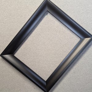 May include: A black picture frame with a beveled edge. The frame is empty and angled, showcasing its clean lines and matte finish. The frame is designed to hold a photograph or artwork.