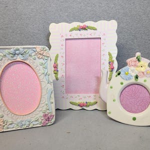 May include: Two rectangular picture frames and one teapot-shaped picture frame. The first frame is oval-centered with pastel floral relief. The second is a scalloped frame with striped patterns. The third is a cream teapot with green polka dots.