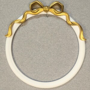 May include: A circular white picture frame featuring a beaded inner edge and a decorative gold-colored ribbon bow accent at the top. The frame has a smooth finish and a classic round shape, designed to hold a circular image or display item.
