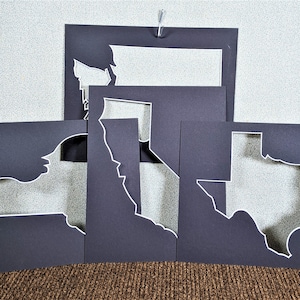 May include: Five black paper mats with white outlines of different US states cut out. The states are Washington, California, New York, Texas, and a smaller state that is not visible.