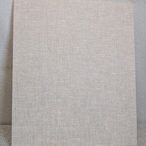 May include: A rectangular board covered in a light beige, woven linen fabric. The material features a visible, uniform cross-hatch texture characteristic of natural flax fibres, presenting a neutral, solid-coloured surface with a matte finish.