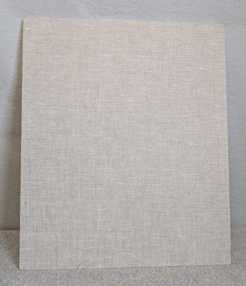May include: A rectangular board covered in a light beige, woven linen fabric. The material features a visible, uniform cross-hatch texture characteristic of natural flax fibres, presenting a neutral, solid-coloured surface with a matte finish.