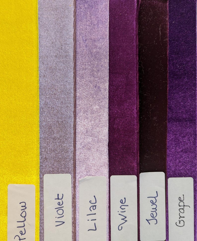 May include: Six vertical strips of velvet ribbon arranged side by side in yellow, violet, lilac, wine, jewel, and grape colors. Each strip features a white rectangular label at the bottom with the corresponding color name written in black ink.