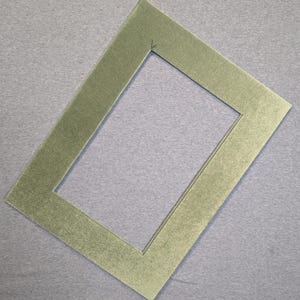 May include: A rectangular picture frame mat with a textured, light olive green surface. The mat features a clean, centered rectangular cutout with sharp, precise edges, designed to frame artwork or photographs with a consistent border width.