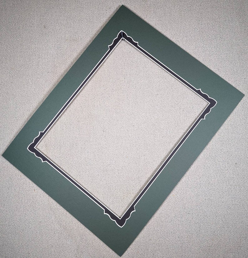 May include: A rectangular dark green picture mat with a textured surface. The inner opening features a decorative scalloped edge highlighted by a thin white border, creating a distinct frame for artwork or photographs within the matting.