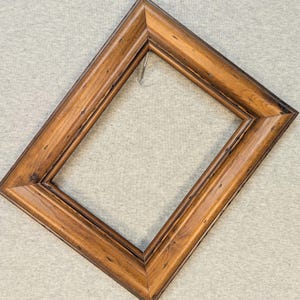 May include: A rectangular picture frame made of medium-toned brown wood with a visible natural grain pattern. The frame is oriented diagonally and features a beveled edge design with dark brown accents along the inner and outer borders.