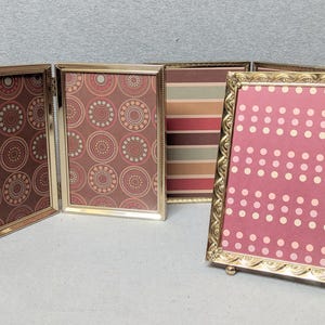 May include: A four-panel folding picture frame with a metallic gold finish. The frames feature decorative edges and contain inserts with various patterns, including brown circles, horizontal stripes, and pink with white polka dots.
