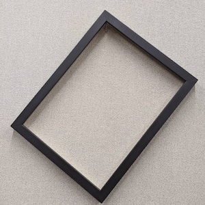 May include: A black picture frame with a simple, modern design. The frame is rectangular and has a matte finish. The frame is empty, ready to display a photo or artwork. The frame is angled on a neutral background.