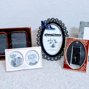 May include: A collection of decorative picture frames in various styles. Frames include a double-opening tortoiseshell frame, a silver frame with two circular openings, an oval frame with a blue shoe accent, a rectangular gray frame, and a red and gold frame.