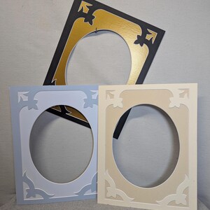 May include: Three decorative picture frames in different colours. One frame is black with a gold interior, another is light blue, and the third is beige. Each frame has an oval opening and decorative corner accents.
