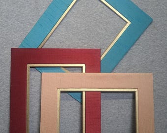 May include: Three decorative picture frames in shades of teal, red, and beige. Each frame has a gold-colored inner border. The frames are rectangular, with one teal frame positioned diagonally. The textures appear to be fabric.