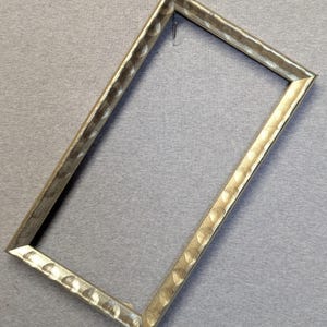 May include: A rectangular picture frame with a textured, hammered metal finish in a light gold color. The frame is empty, with a simple hook for hanging. The frame's design is modern and minimalist.